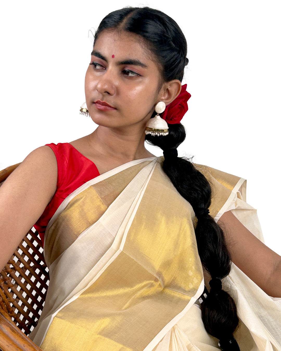 Woman wearing a traditional kerala kasavu saree with a white and gold design, sitting on a woven chair.