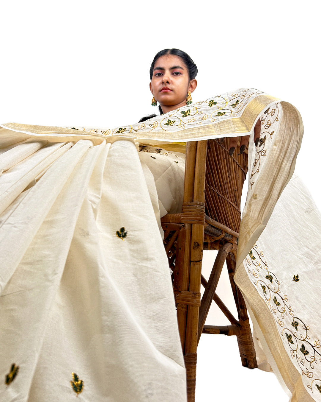 Woman sitting on a woven chair wearing a white saree with gold embroidery.