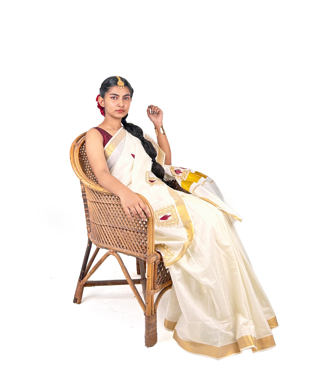 Woman in a white saree with gold border sitting on a wicker chair against a white background